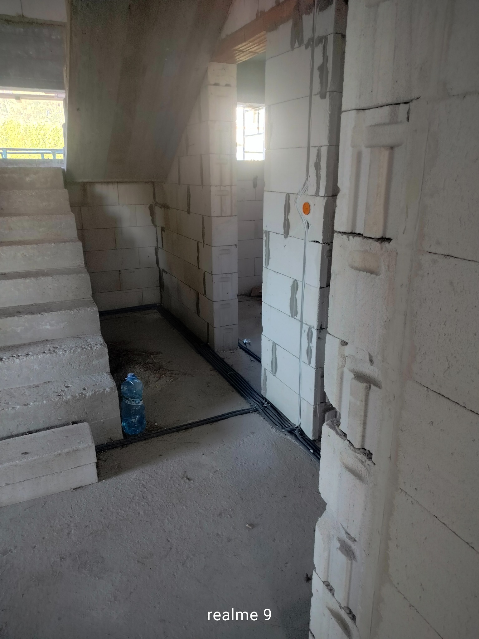 Wnętrze surowego domu w trakcie budowy, widoczne ściany z bloczków, schody, instalacja elektryczna w peszlach przy podłodze i puszka elektryczna na ścianie. W tle okno i butelka wody mineralnej.