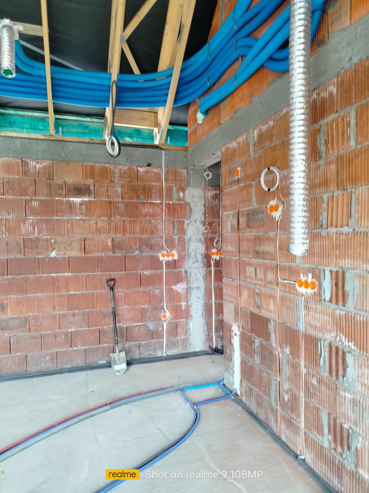 Wnętrze budynku w trakcie prac instalacyjnych: widoczne przewody elektryczne w ścianie z cegły, niebieskie rury wentylacyjne pod sufitem i łopata oparta o ścianę.