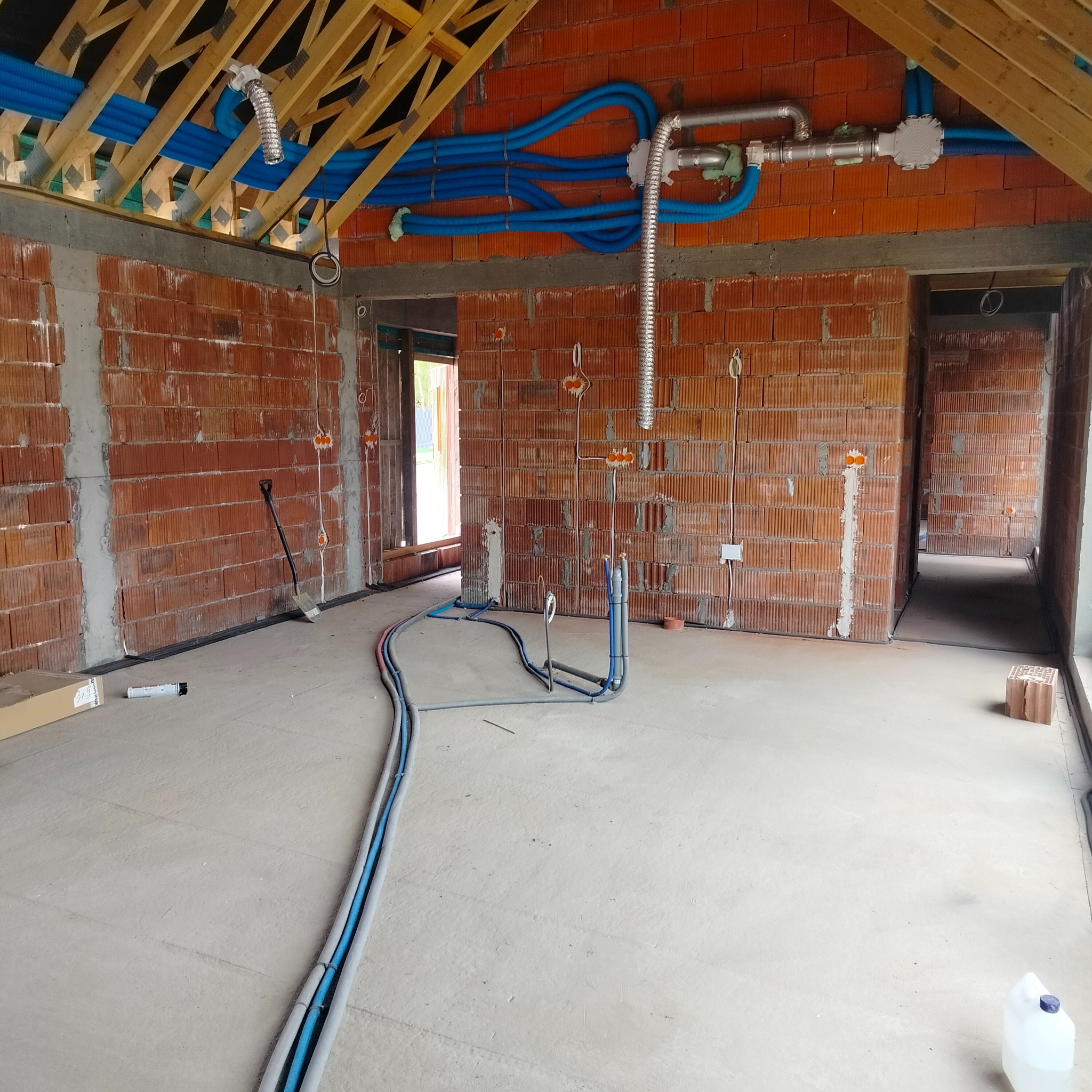 Wnętrze w trakcie budowy z widocznymi przewodami instalacji elektrycznej i wentylacyjnej, kable ułożone na podłodze, ściany z czerwonej cegły, drewniana konstrukcja dachu.