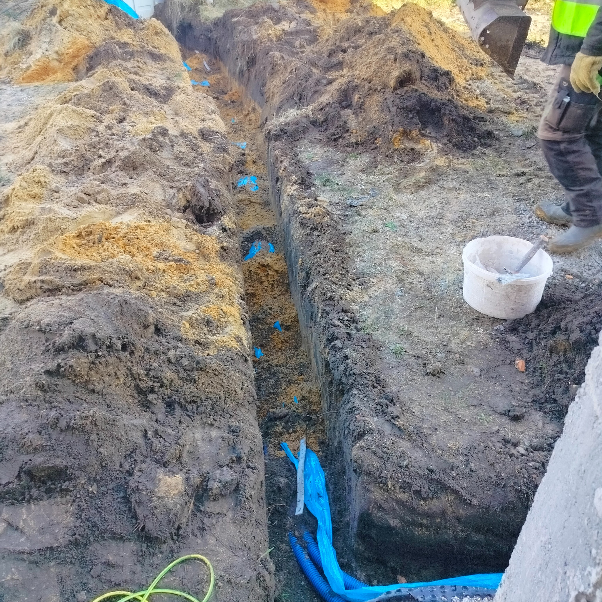 Wykop w ziemi z ułożonymi niebieskimi rurami karbowanymi i niebieską folią ochronną, widoczny fragment koparki i pracownik w odblaskowej kamizelce.