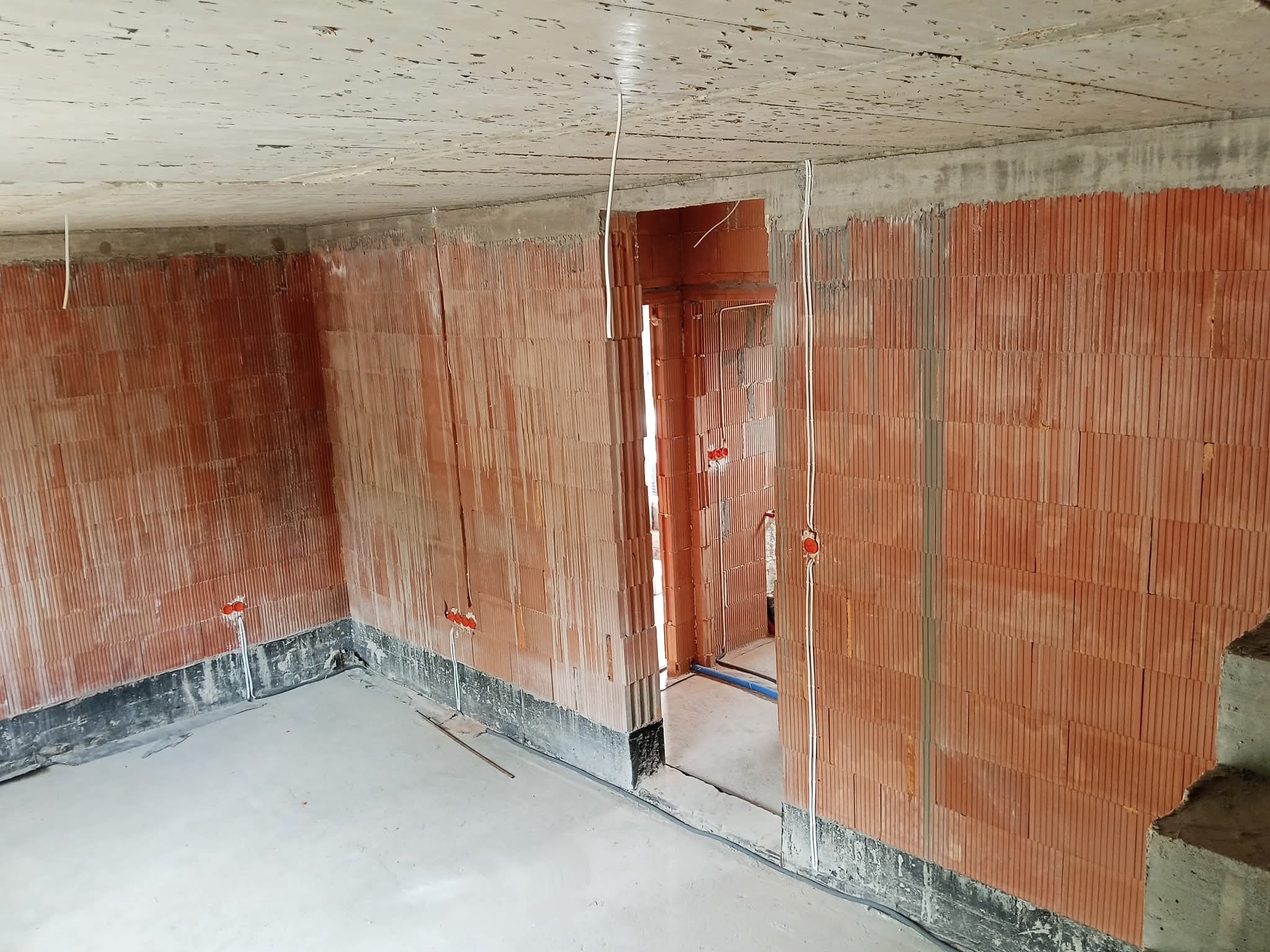 Surowy stan: Instalacja elektryczna wewnątrz budynku z czerwonej cegły. Widoczne kable i puszki instalacyjne. Podłoga betonowa, sufit surowy. W tle otwór drzwiowy.