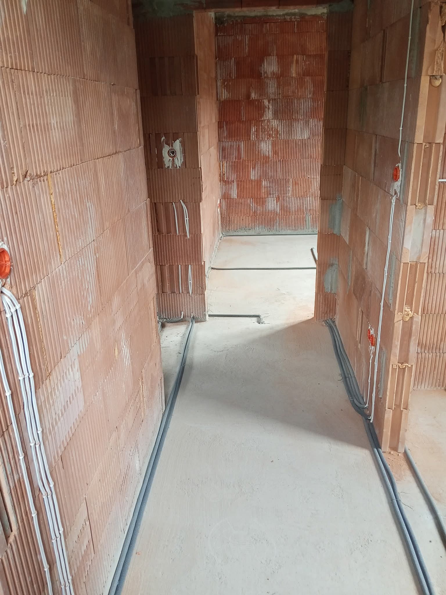 Wnętrze w trakcie instalacji elektrycznej: widoczne kable w ścianach z cegły dziurawki oraz peszle na podłodze. Pomarańczowe puszki elektryczne na ścianach.