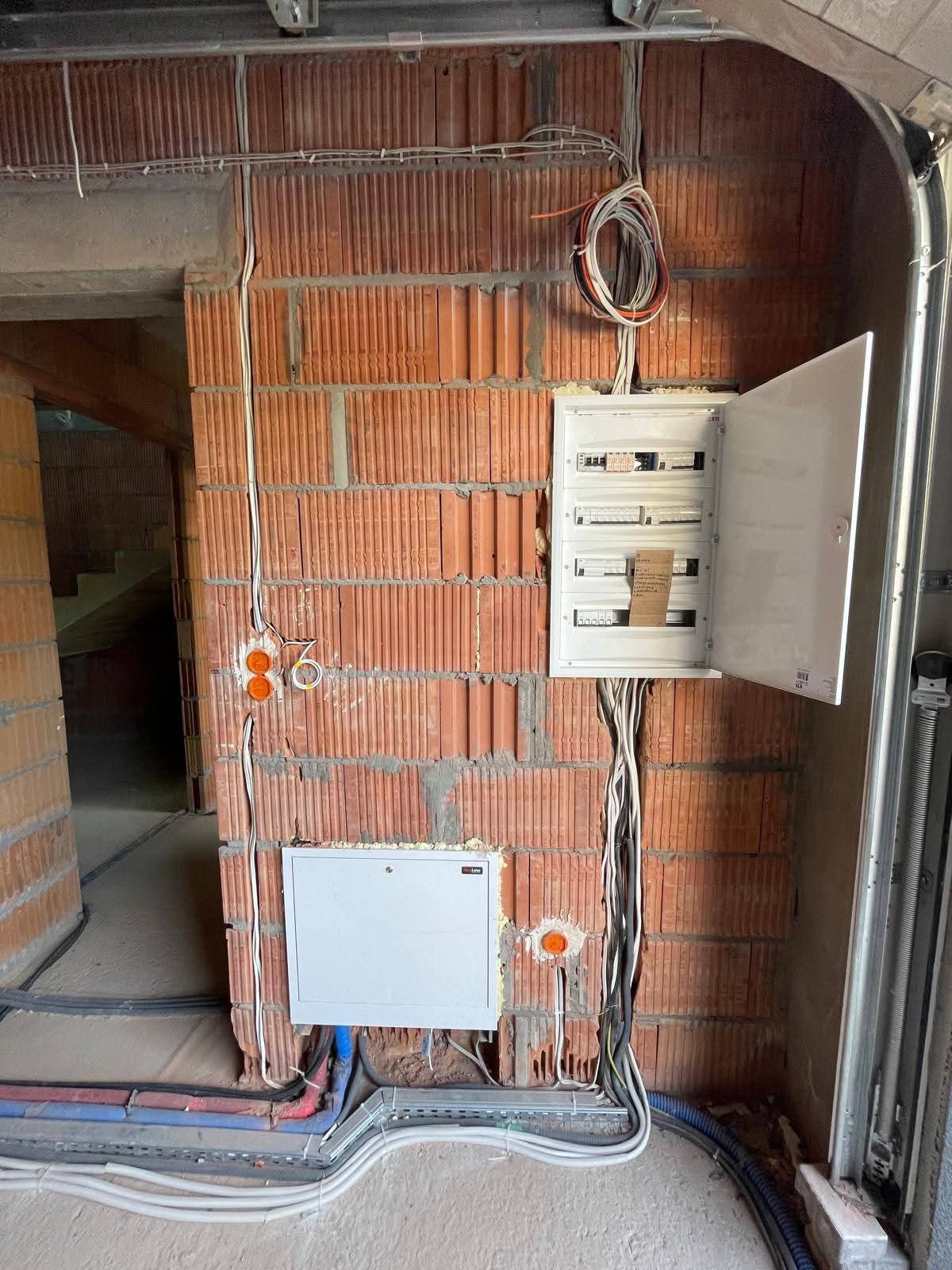 Rozdzielnica elektryczna z otwartymi drzwiczkami na ceglanej ścianie w trakcie montażu. Widoczne przewody i puszki instalacyjne. Surowy stan wykończenia wnętrza.