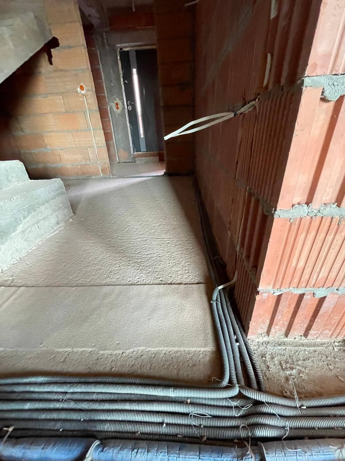 Kable elektryczne w peszlach ułożone wzdłuż ściany z czerwonej cegły w surowym wnętrzu budynku w trakcie budowy. Widoczne puszki elektryczne w ścianie.