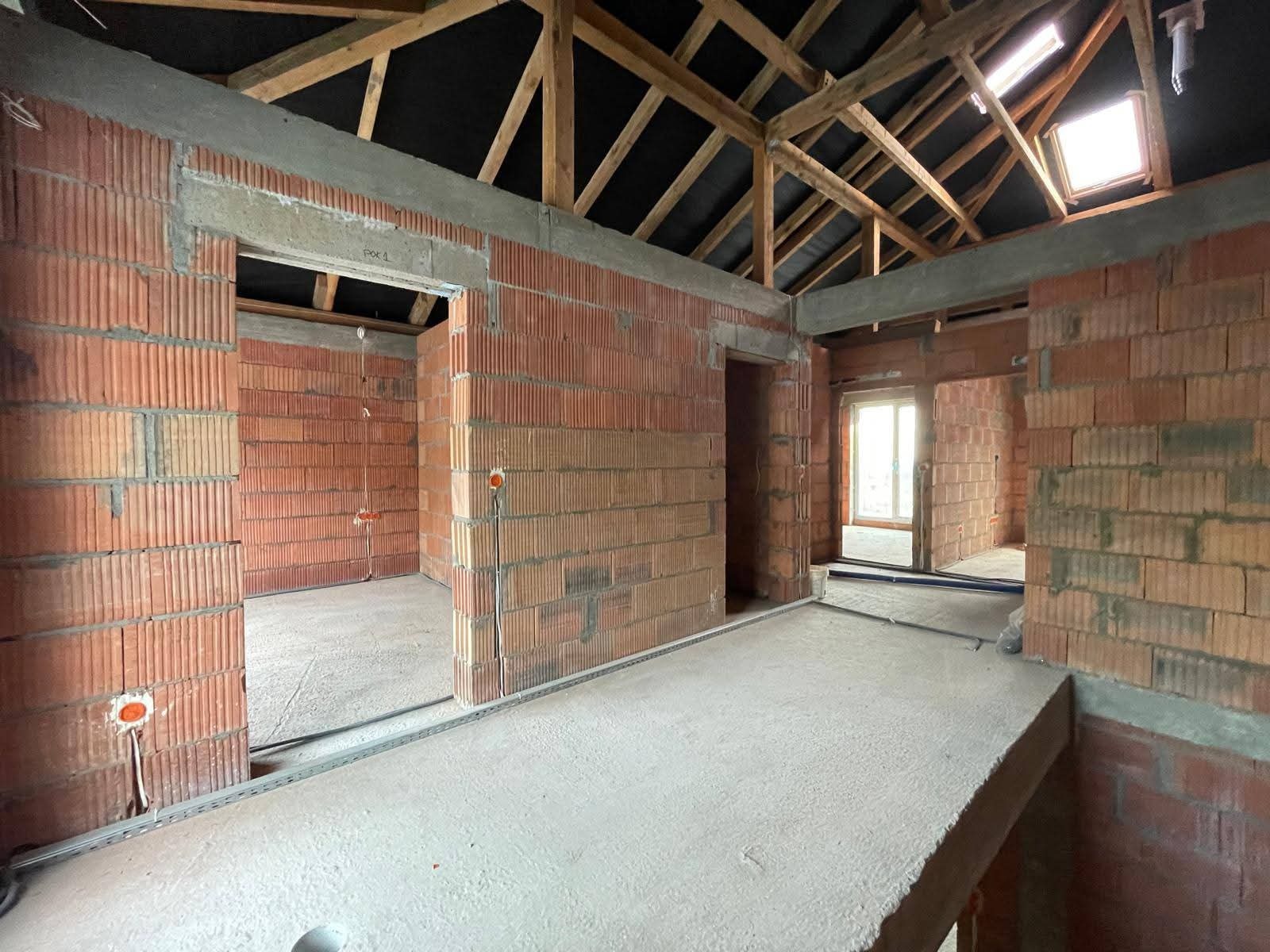 Surowy stan wnętrza domu z czerwonej cegły z widocznymi puszkami elektrycznymi i drewnianą konstrukcją dachu. Okno dachowe wpuszcza światło do środka.