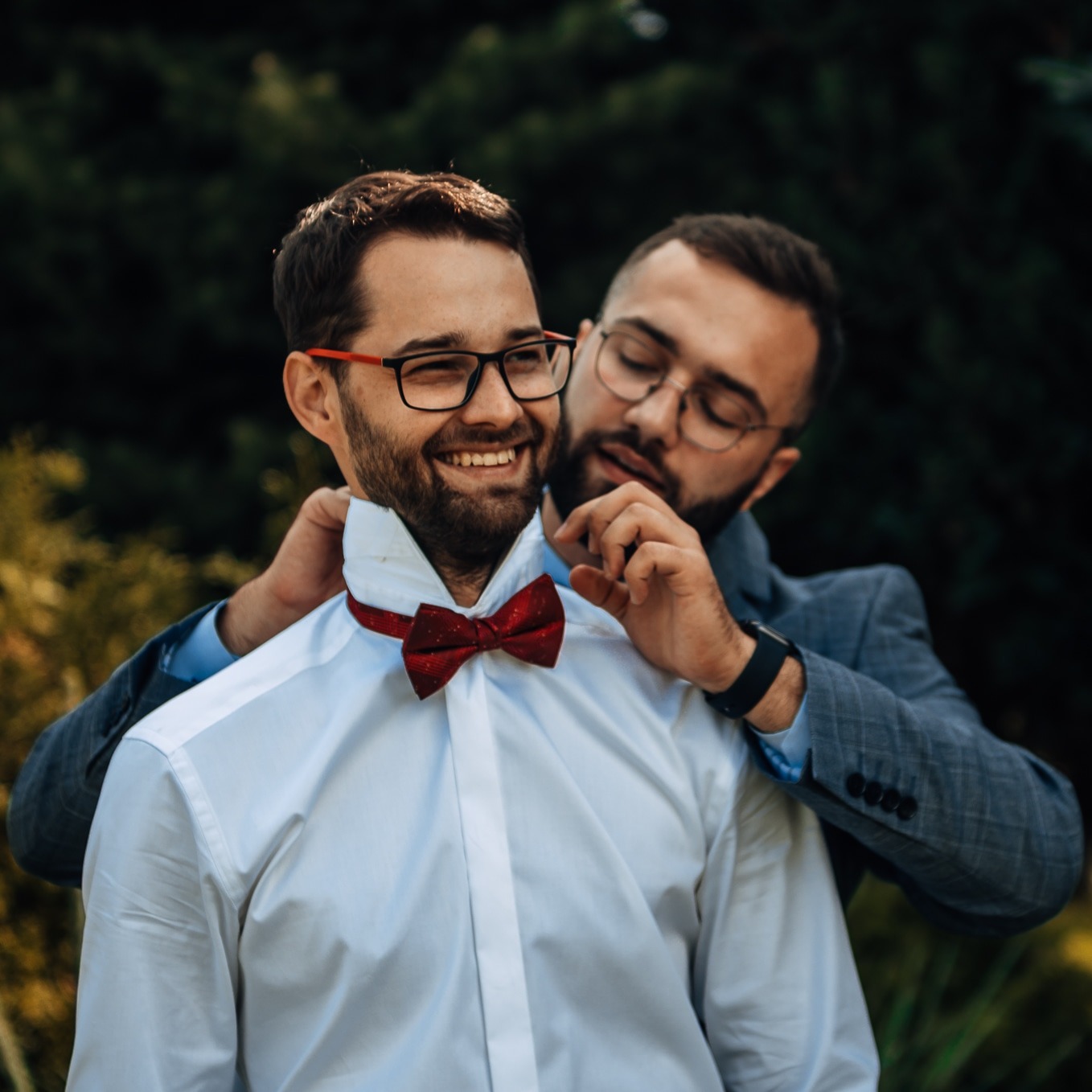 Mężczyzna poprawia muszkę drugiemu mężczyźnie, obaj uśmiechnięci, na tle zieleni.