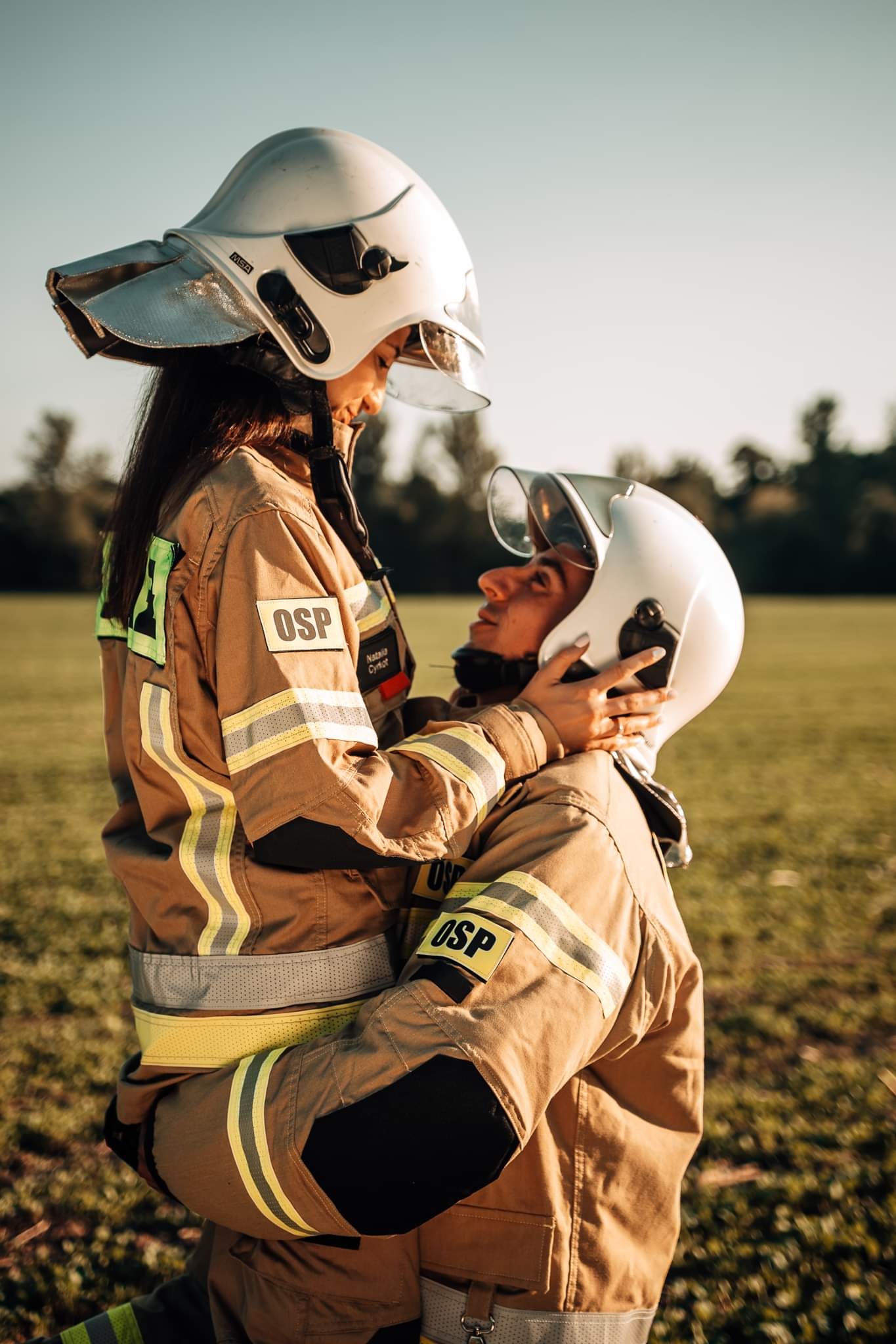 Para ubrana w stroje strażackie i hełmy spogląda na siebie z czułością na tle zielonej łąki.