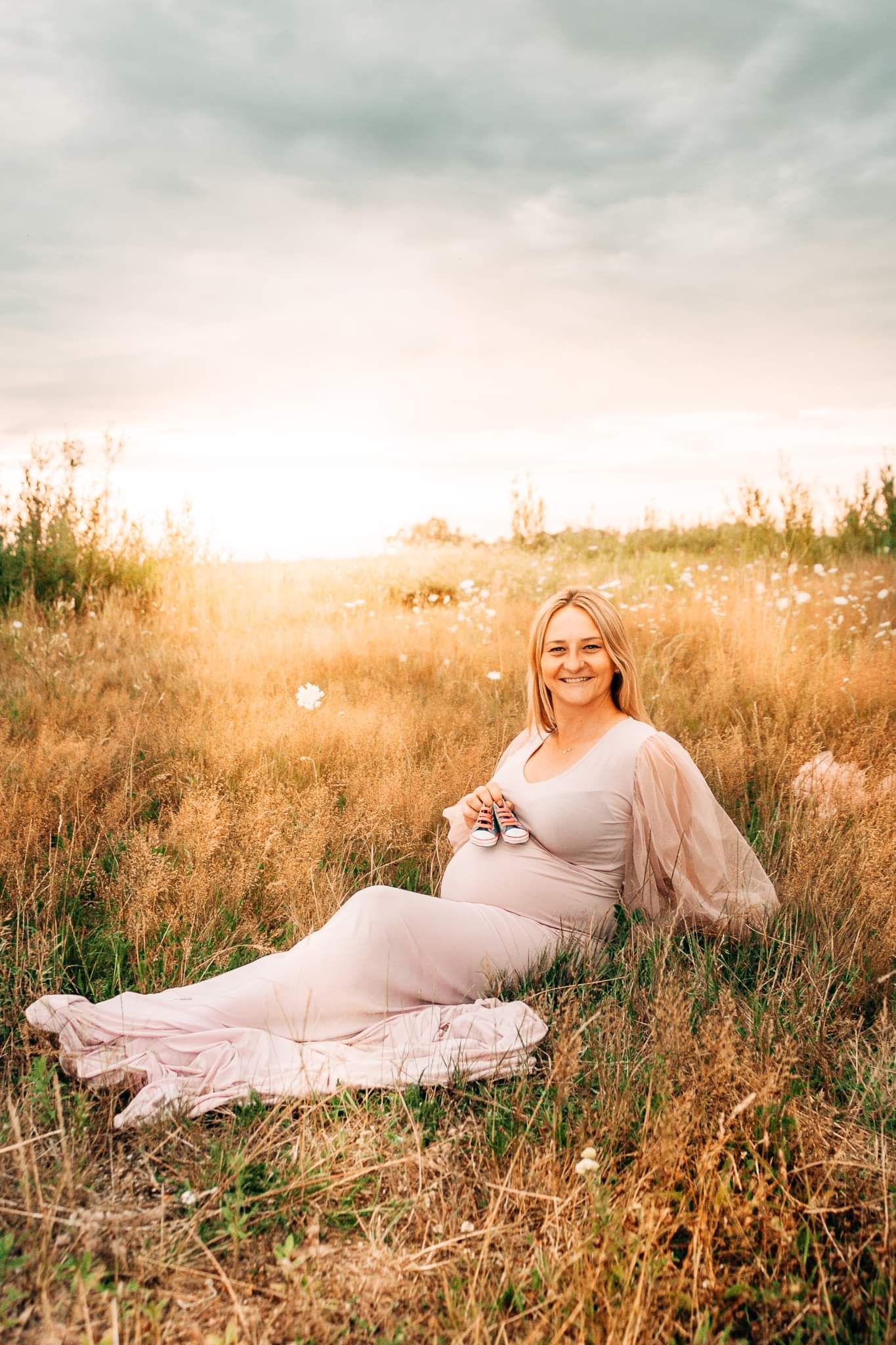 Uśmiechnięta kobieta w zwiewnej, pudrowej sukni ciążowej, siedząca w wysokiej trawie z małymi, czerwono-białymi bucikami w dłoniach, w tle rozmyte, letnie pole o zachodzie słońca.