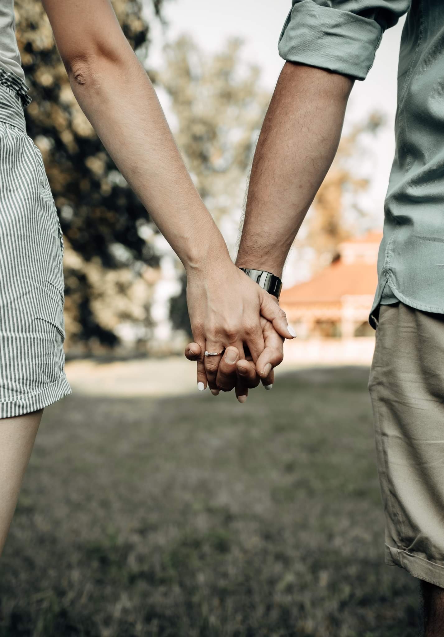 Złączone dłonie pary, widoczny pierścionek zaręczynowy na palcu kobiety, w tle zieleń i budynek z czerwoną dachówką.