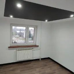 Pokój po remoncie: brązowe panele podłogowe, białe ściany, okno z drewnianym parapetem, biały grzejnik i podwieszany sufit z czarną wstawką i punktowym oświetleniem.