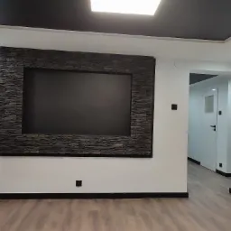 Salon z czarnym sufitem, drewnianą podłogą i ścianą z kamienną ramą wokół czarnego prostokąta, widok na korytarz z białymi drzwiami.