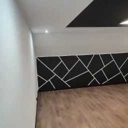 Pomieszczenie z drewnianą podłogą, białą ścianą i fragmentem ściany pomalowanej na czarno z białymi, geometrycznymi wzorami, oraz czarnym fragmentem sufitu.