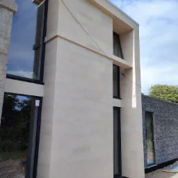 Nowoczesny budynek z elewacją z jasnego kamienia i dużymi oknami z czarnymi ramami, otoczony terenem w trakcie prac wykończeniowych.