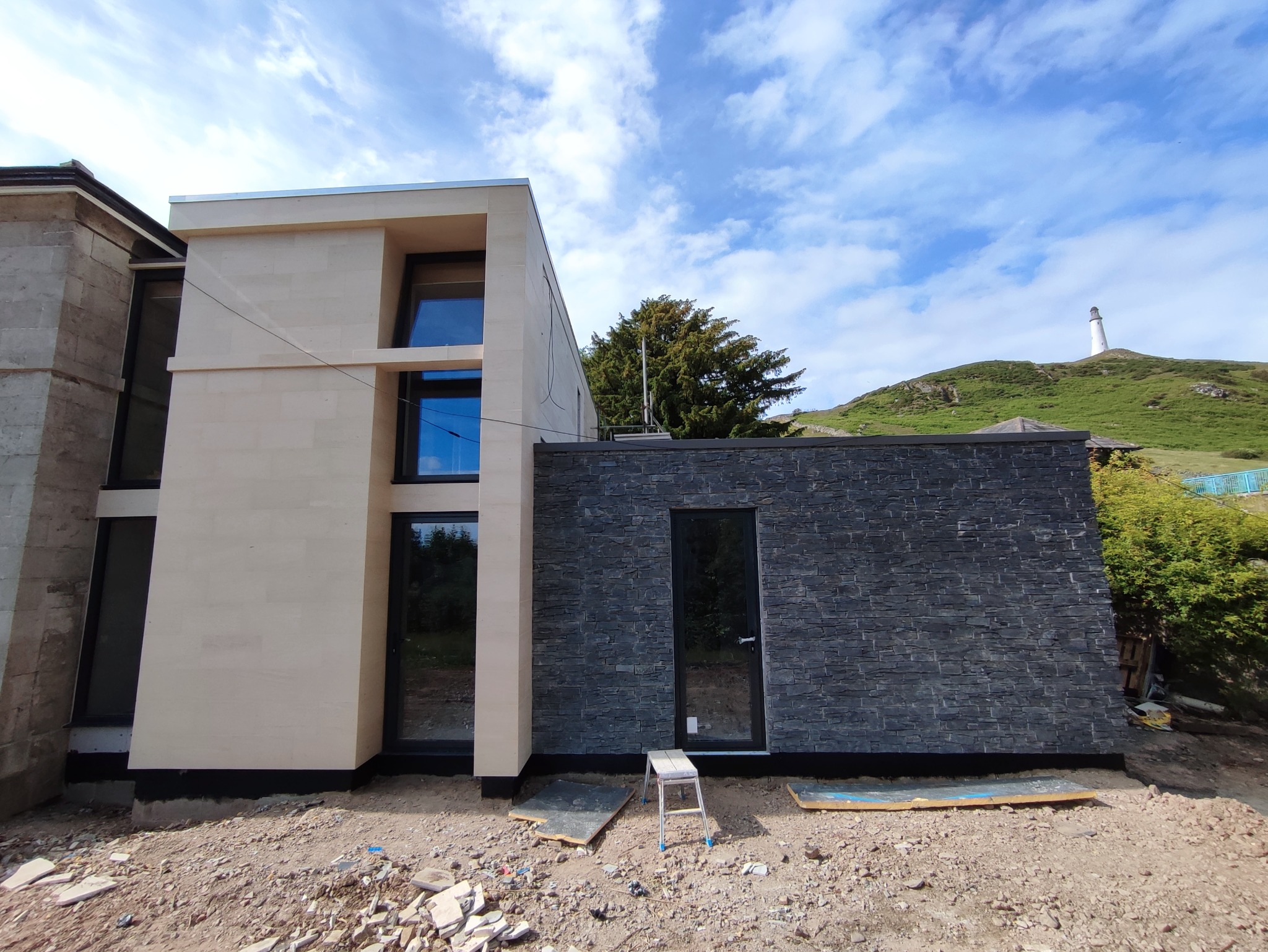 Nowoczesny dom z elewacją z jasnego kamienia i ciemnego łupka, na tle wzgórza z latarnią morską, widoczne ślady prac wykończeniowych.