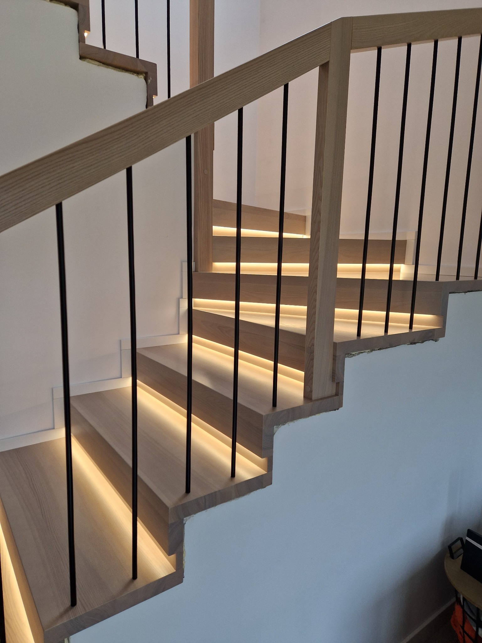 Nowoczesne schody z jasnego drewna z podświetleniem LED i czarnymi, metalowymi słupkami balustrady. Minimalistyczny design, ciepłe światło.