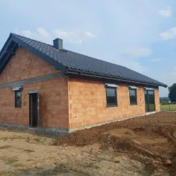 Dom jednorodzinny w trakcie budowy, z ciemnoszarą dachówką ceramiczną, widoczne mury z czerwonej cegły i przygotowany teren.