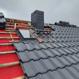 Montaż czarnej dachówki ceramicznej na dachu z oknem dachowym i kominem, widoczna membrana dachowa i narzędzia.