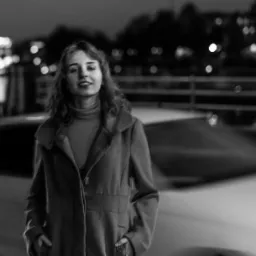 Czarno-białe zdjęcie kobiety w płaszczu na tle nocnego miasta z rozmytym kołem diabelskim i przejeżdżającym samochodem.