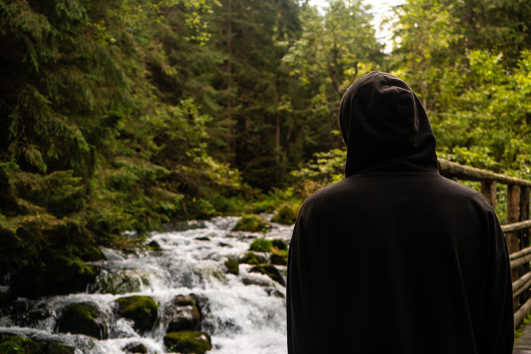 Osoba w czarnej bluzie z kapturem stoi tyłem, obserwując górski potok z kaskadami i bujną zielenią drzew w tle.