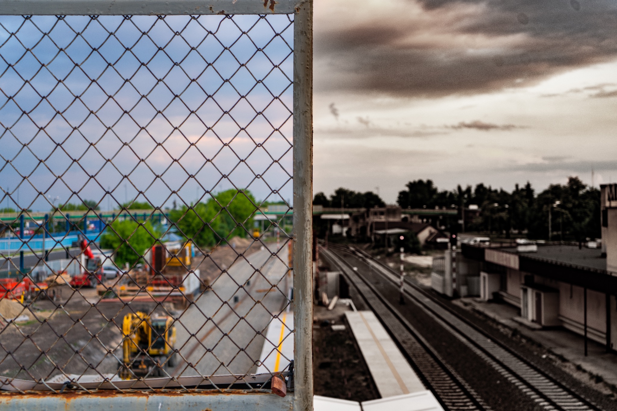 Widok przez zardzewiałą siatkę ogrodzeniową na plac budowy z maszynami i oddaloną stację kolejową w pochmurny dzień.