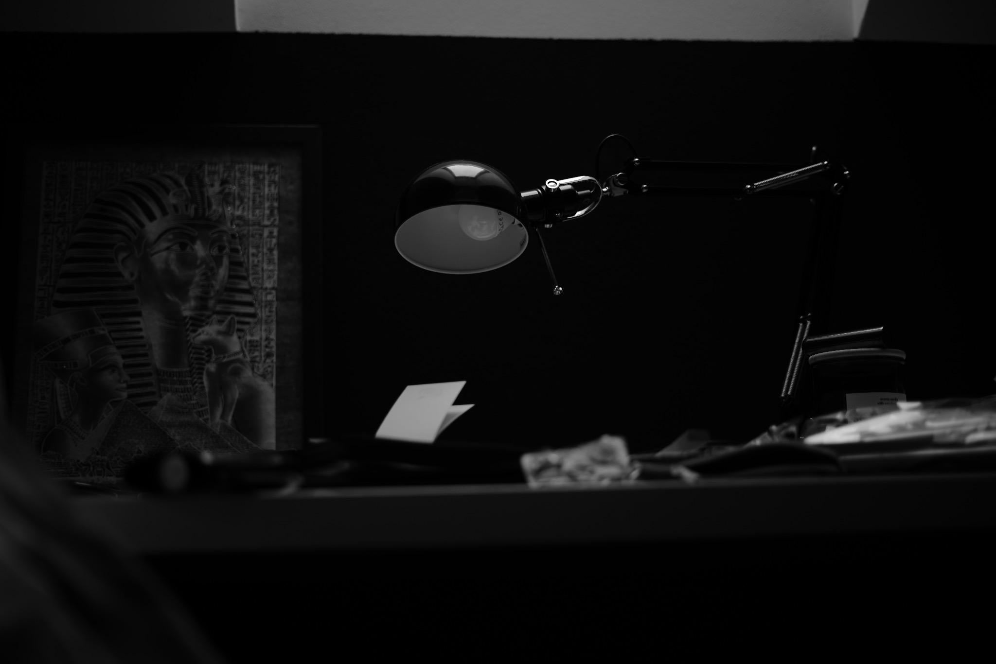 Czarno-białe zdjęcie biurka z lampką kreślarską oświetlającą notatki, w tle oprawiony obraz z motywem egipskim.