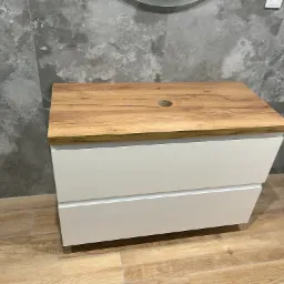 Biała szafka łazienkowa z drewnianym blatem z otworem na umywalkę, na tle szarej ściany z płytek imitujących kamień i drewnianej podłogi, z butelką płynu do prania w rogu.