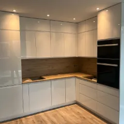 Nowoczesna, minimalistyczna kuchnia z białymi, lakierowanymi frontami, drewnianym blatem i fornirem na ścianie, oświetleniem punktowym i wbudowanym piekarnikiem.