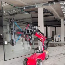 Czerwony robot do montażu szyb podnosi dużą taflę szkła w surowym wnętrzu budynku w trakcie budowy.