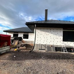 Owsiak Patryk Okna - Dom jednorodzinny w trakcie ocieplania, z zamontowanymi roletami zewnętrznymi, widoczne rusztowania, styropian z czarnymi kołkami, przyczepa z materiałami budowlanymi.