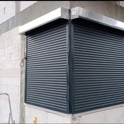 Owsiak Patryk Okna - Montaż ciemnych rolet zewnętrznych na rogu budynku z białej cegły, widoczny fragment drabiny i poziomicy.