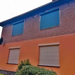 Owsiak Patryk Okna - Dwukondygnacyjny dom z oknami zabezpieczonymi roletami zewnętrznymi: niebieskie na górze i beżowe na dole, z widoczną anteną satelitarną na elewacji.