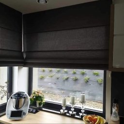 Owsiak Patryk Okna - Ciemne, tekstylne rolety rzymskie w kuchni, widok na zewnętrzny ogród z niską roślinnością płożącą się po ścianie, na blacie kuchennym robot kuchenny, świeczniki i owoce.
