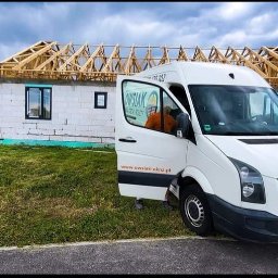 Owsiak Patryk Okna - Montaż okna w surowym budynku z widocznym szkieletem dachu, obok zaparkowany biały bus firmy z otwartymi drzwiami.