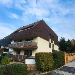 Owsiak Patryk Okna - Dwupiętrowy budynek mieszkalny z balkonami, brązowym dachem i jasną elewacją, otoczony zielenią i niskim ceglanym murkiem, widziany z poziomu ulicy w słoneczny dzień.