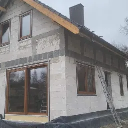 Surowy stan domu w trakcie budowy z zamontowanymi oknami w drewnianych ramach, widoczna drabina oparta o ścianę i fragment dachu.