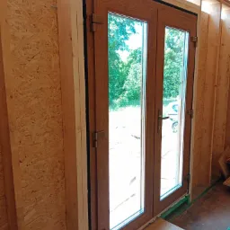 Montaż dwuskrzydłowych drzwi balkonowych z brązową ramą w surowym stanie budynku, widoczne płyty OSB na ścianach i fragment zielonego narzędzia u dołu.