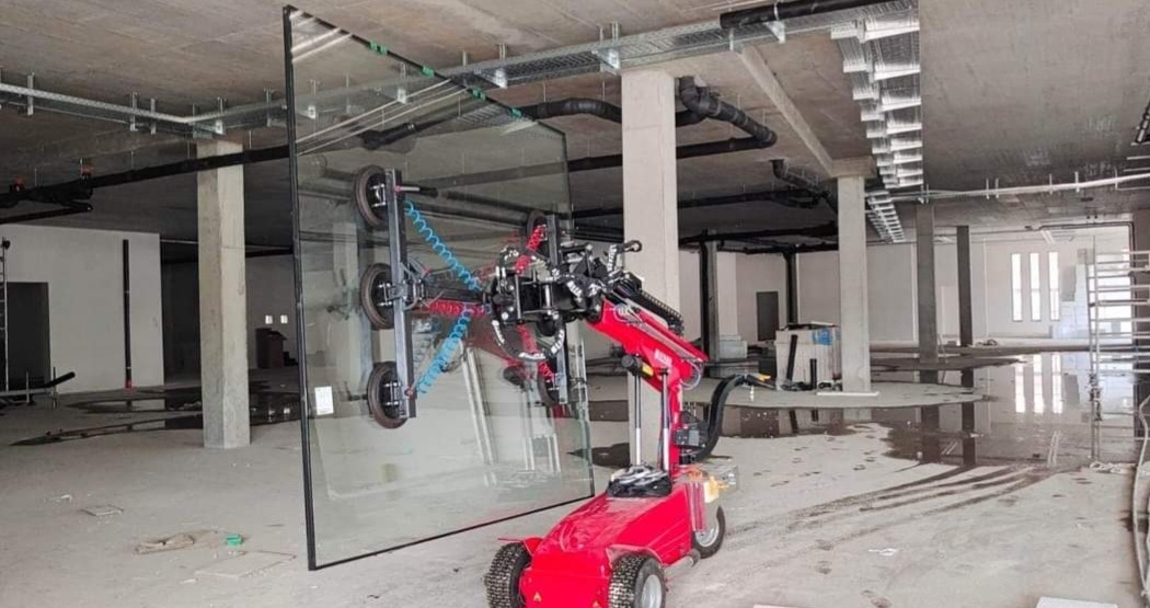 Czerwony robot do montażu szyb podnosi dużą taflę szkła w surowym wnętrzu budynku w trakcie budowy.