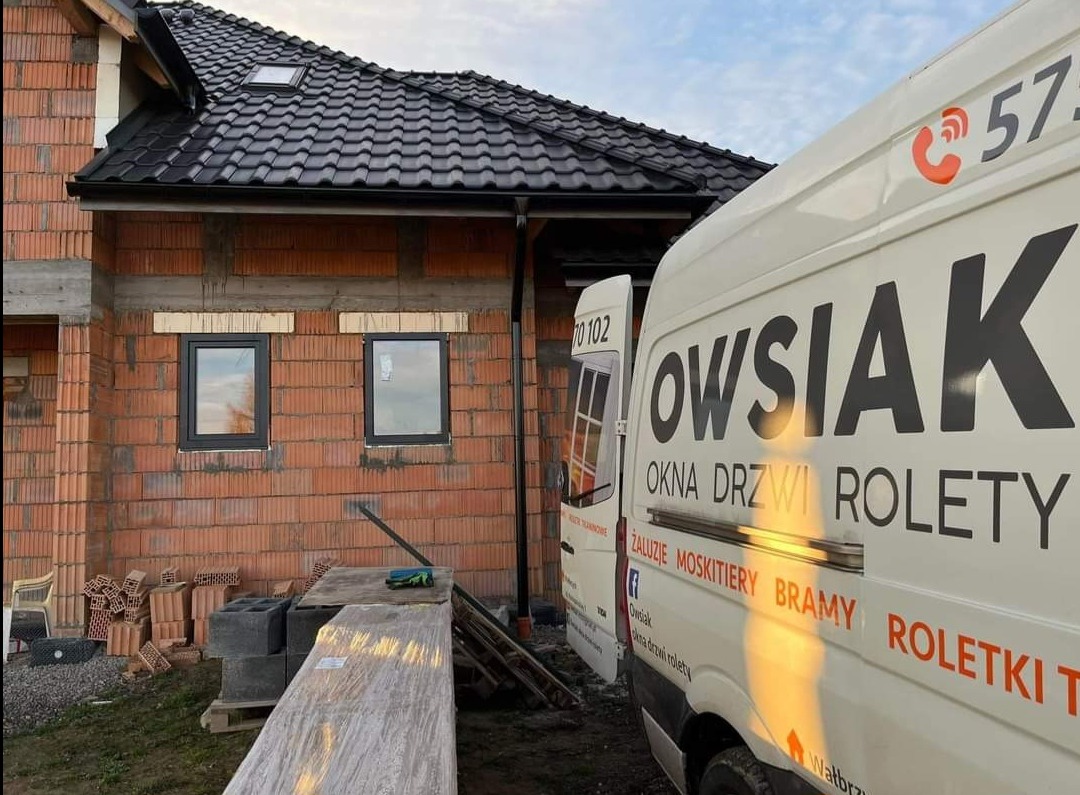 Nowy dom w trakcie budowy z zamontowanymi czarnymi oknami PCV, na pierwszym planie częściowo widoczny biały samochód dostawczy firmy Owsziak z reklamą okien, drzwi i rolet.