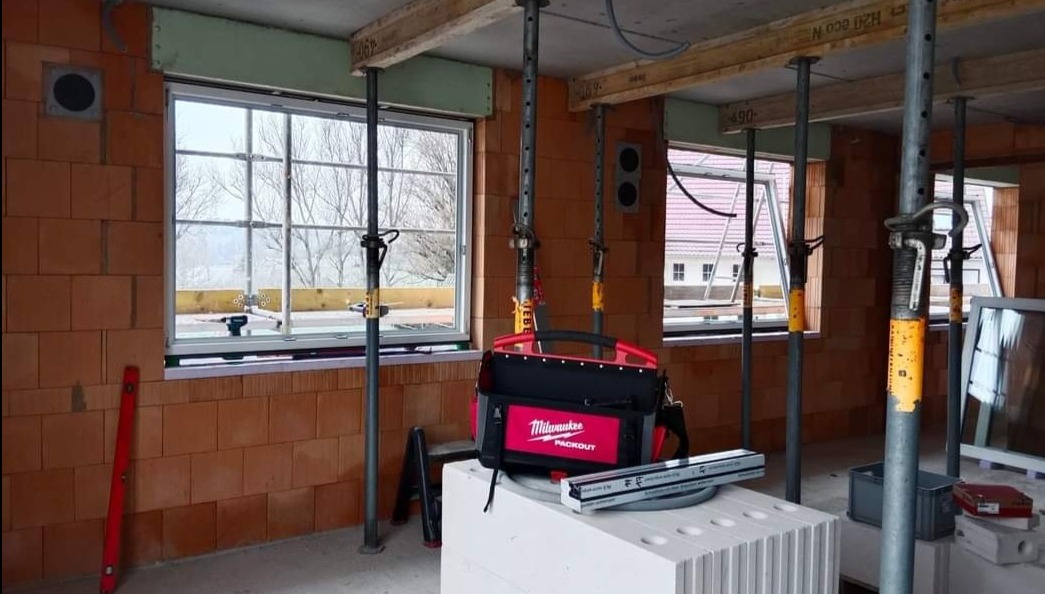 Wnętrze budynku w trakcie budowy, widoczne okna z białymi ramami, podpory stropowe, poziomica i torba narzędziowa Milwaukee Packout na bloczkach.