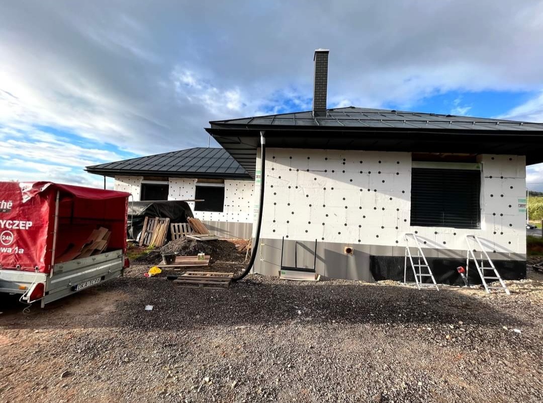 Dom jednorodzinny w trakcie ocieplania, z zamontowanymi roletami zewnętrznymi, widoczne rusztowania, styropian z czarnymi kołkami, przyczepa z materiałami budowlanymi.