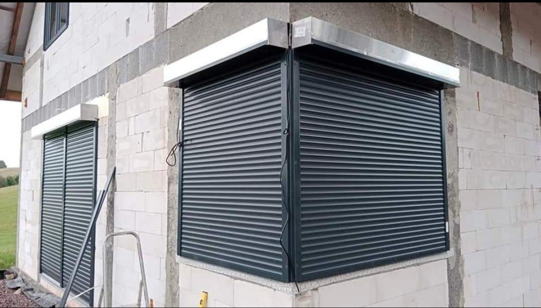 Montaż ciemnych rolet zewnętrznych na rogu budynku z białej cegły, widoczny fragment drabiny i poziomicy.