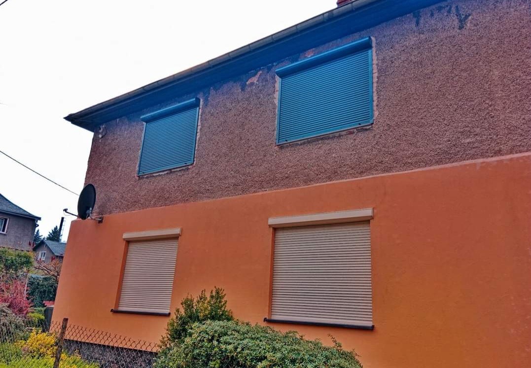 Dwukondygnacyjny dom z oknami zabezpieczonymi roletami zewnętrznymi: niebieskie na górze i beżowe na dole, z widoczną anteną satelitarną na elewacji.