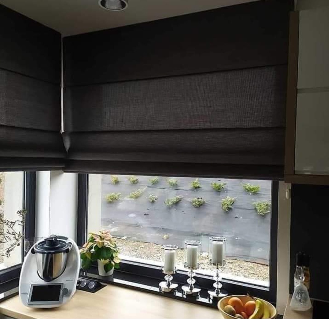 Ciemne, tekstylne rolety rzymskie w kuchni, widok na zewnętrzny ogród z niską roślinnością płożącą się po ścianie, na blacie kuchennym robot kuchenny, świeczniki i owoce.