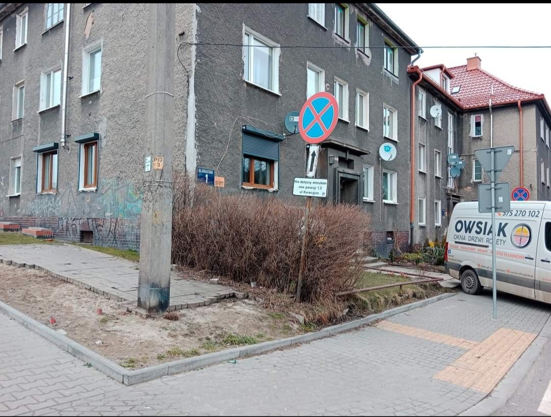 Szary blok mieszkalny z oknami PCV i roletami zewnętrznymi, częściowo zasłonięty krzewami, z zaparkowanym białym samochodem dostawczym firmy Owsik oferującej okna, drzwi i rolety.