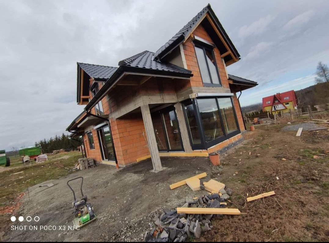 Surowy budynek w trakcie budowy z zamontowanymi oknami i roletami zewnętrznymi, widoczna zagęszczarka na nierównym terenie.