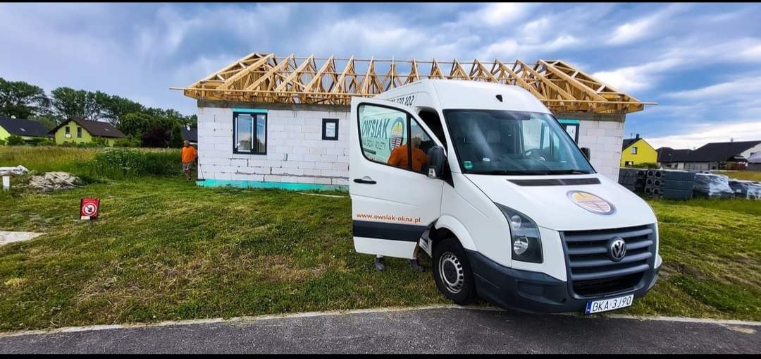 Montaż okna w surowym budynku z widocznym szkieletem dachu, obok zaparkowany biały bus firmy z otwartymi drzwiami.