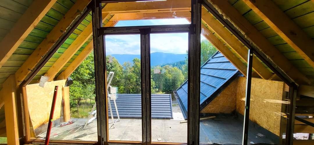 Montaż okien w surowym stanie poddasza z widokiem na góry, z poziomnicą i drabiną opartą o ramę okienną.