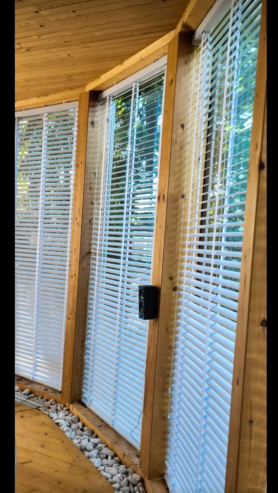 Białe żaluzje poziome zamontowane w drewnianej konstrukcji okiennej, widoczne z perspektywy podłogi wyłożonej drewnem i kamieniami.
