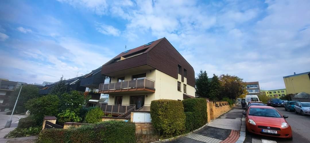 Dwupiętrowy budynek mieszkalny z balkonami, brązowym dachem i jasną elewacją, otoczony zielenią i niskim ceglanym murkiem, widziany z poziomu ulicy w słoneczny dzień.