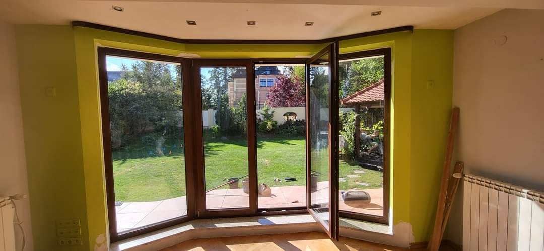 Montaż okien z brązowymi ramami w narożnym pokoju z zielonymi ścianami, widok na ogród przez otwarte okno.